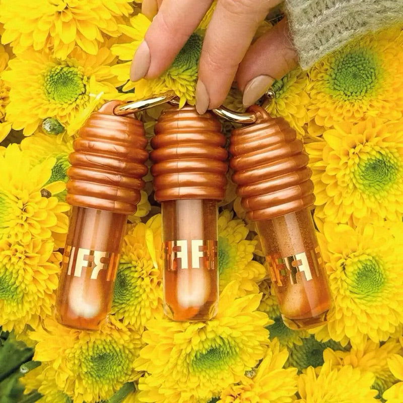 Três batons dourados em formato de cápsula, segurados por uma mão, sobre flores amarelas