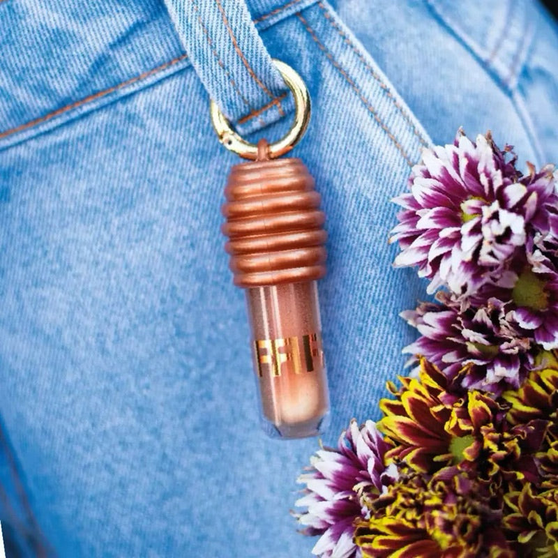 Batom dourado pendurado em calça jeans com flores roxas ao lado, cosmético em destaque.